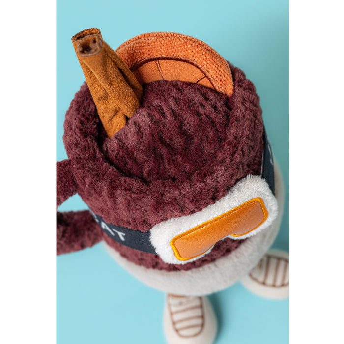 aerial view of a plush soft toy in the shape of a mug of mulled hot drink with ski goggles and festive toppings. The mug is against a blue backdrop. - Image