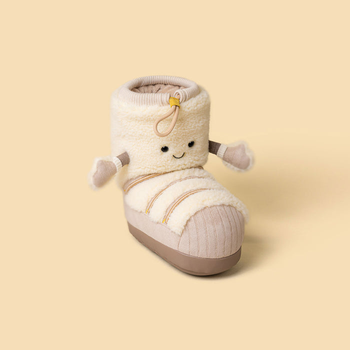 A plush soft toy in the shape of a ski boot in cream and beige with a cute embroidered face and small fabric arms, shown on a solid pale yellow background. - Image