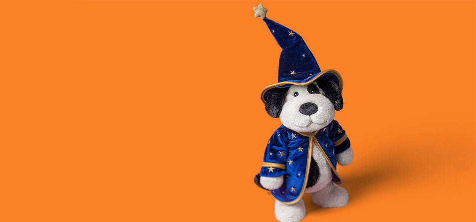 A plush dog toy dressed as a wizard, wearing a blue robe and pointed hat with gold stars, standing against a solid orange background. - Image