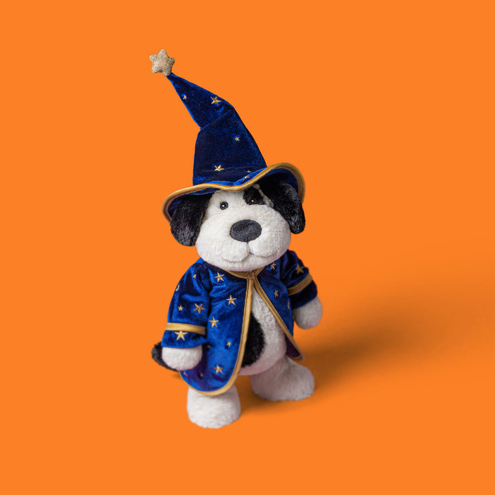 A plush dog toy dressed as a wizard, wearing a blue velvet hat and matching robe with gold star details, photographed against a solid orange background. - Image