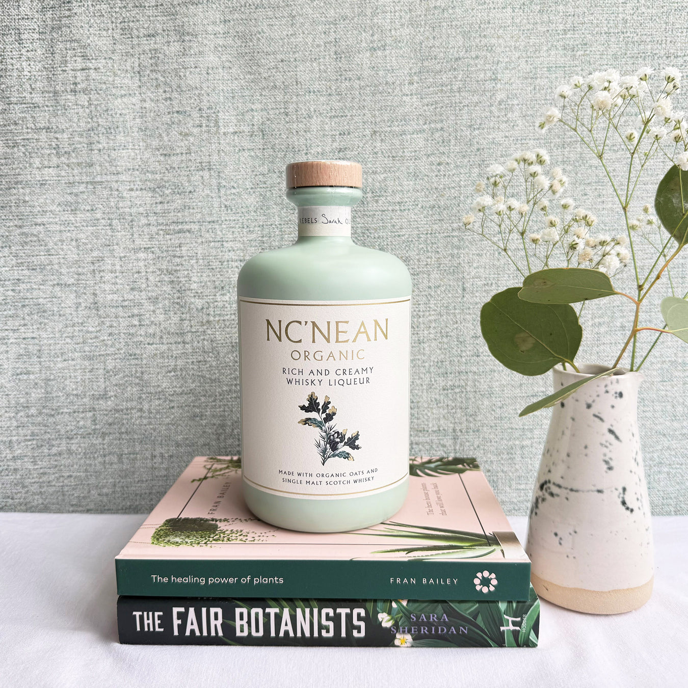 Bottle of NC'NEAN Organic whisky liqueur on books with a vase of flowers against a gray textured wall. - Image
