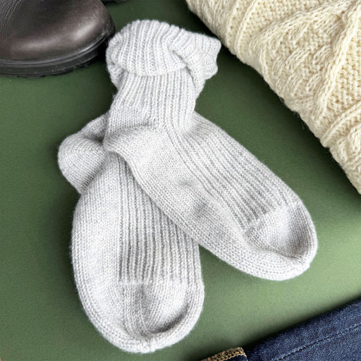 A pair of light grey ribbed knit ankle socks laid flat on a green surface, styled with a cream cable-knit sweater, a dark boot, and blue jeans. - Image