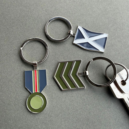 Four metal keychains with enamel military-style insignia are arranged on a grey surface. The set includes a round silver keyring with a blue, white, and red ribbon medallion, a silver keyring with a blue and white saltire flag, a silver keyring with green chevron stripes, and a partial silver keyring with a matching key. - Image
