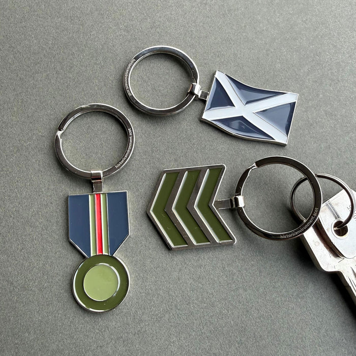 Four metal keychains with enamel military-style insignia are arranged on a grey surface. The set includes a round silver keyring with a blue, white, and red ribbon medallion, a silver keyring with a blue and white saltire flag, a silver keyring with green chevron stripes, and a partial silver keyring with a matching key. - Image