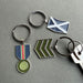 Four metal keychains with enamel military-style insignia are arranged on a grey surface. The set includes a round silver keyring with a blue, white, and red ribbon medallion, a silver keyring with a blue and white saltire flag, a silver keyring with green chevron stripes, and a partial silver keyring with a matching key. - Image