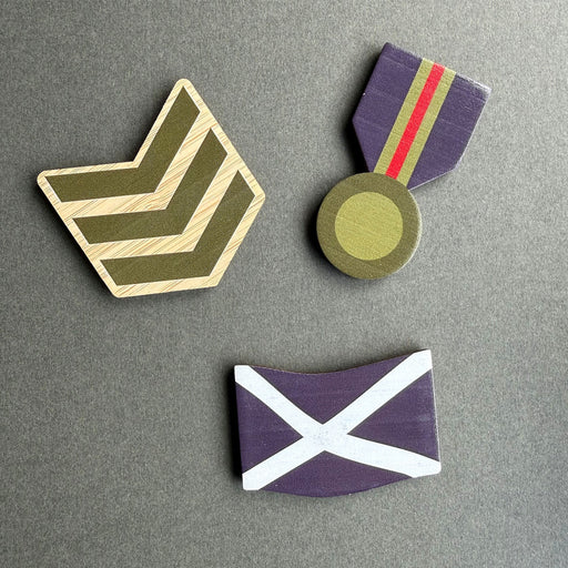 Three wooden military-style magnets arranged on a dark gray background: a chevron rank in olive green and beige, a medal with purple ribbon and gold-toned medallion, and a blue Saltire. - Image