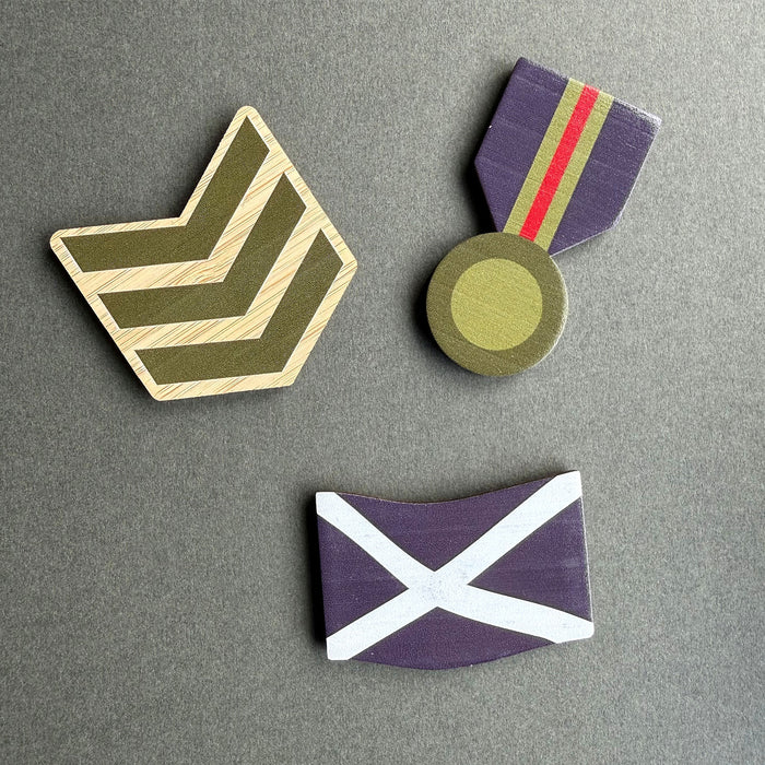 Three wooden military-style magnets arranged on a dark gray background: a chevron rank in olive green and beige, a medal with purple ribbon and gold-toned medallion, and a blue Saltire. - Image
