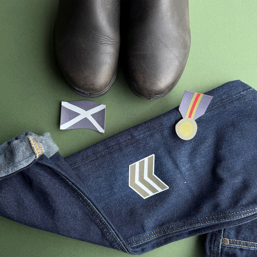 A styled flat-lay of a pair of dark blue denim jeans with embroidered military-style insignia patches, alongside a pair of black boots, a small flag patch,  all arranged on a solid green background. - Image