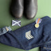 A styled flat-lay of a pair of dark blue denim jeans with embroidered military-style insignia patches, alongside a pair of black boots, a small flag patch,  all arranged on a solid green background. - Image