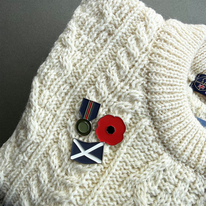 A close-up of a cream cable-knit sweater with several enamel lapel pins pinned to the front, including a red poppy, a blue-and-white saltire flag pin, and other small military-style pins. - Image