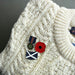 A close-up of a cream cable-knit sweater with several enamel lapel pins pinned to the front, including a red poppy, a blue-and-white saltire flag pin, and other small military-style pins. - Image