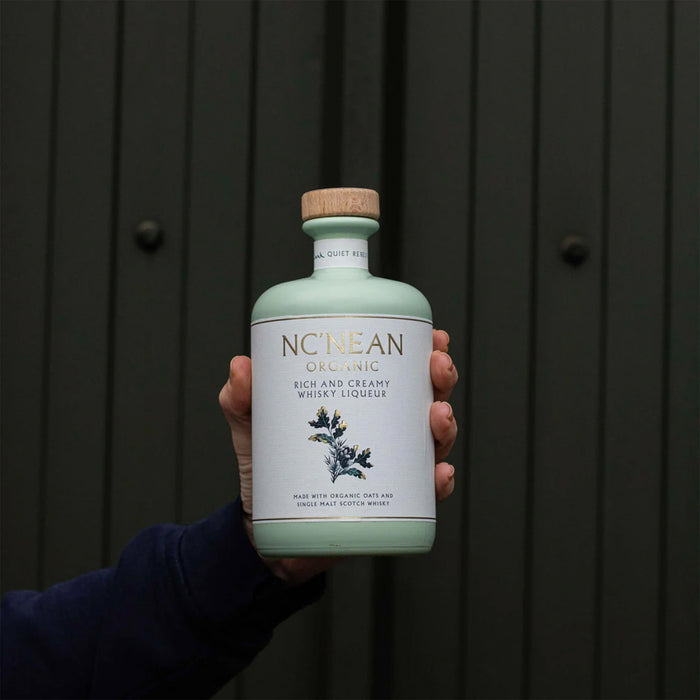 A hand holds a bottle of NcNean Whisky Liqueur against a green door. - Image
