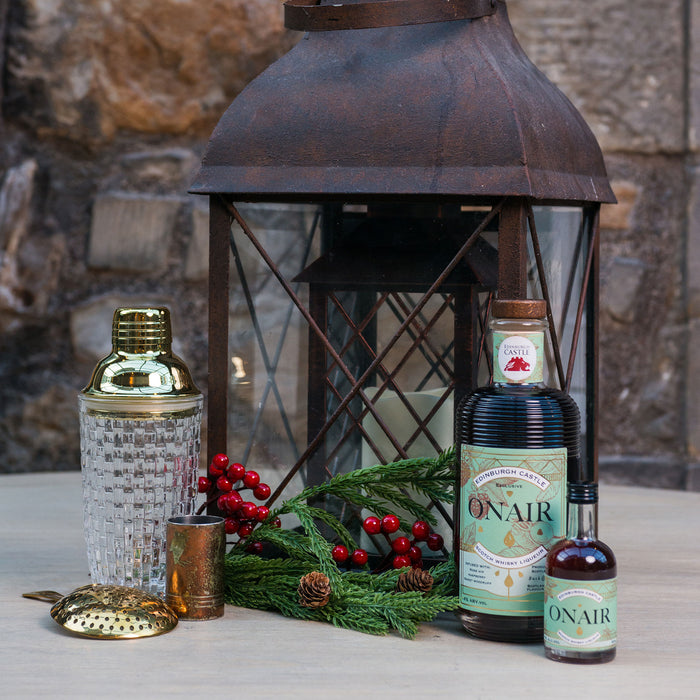 A styled tabletop scene with a large dark metal lantern centered behind several bottles, including a prominent ONAIR bottle and a smaller matching bottle, alongside a faceted glass bottle, gold-toned accessories, evergreen branches, pinecones, and red berries against a stone wall background. - Image