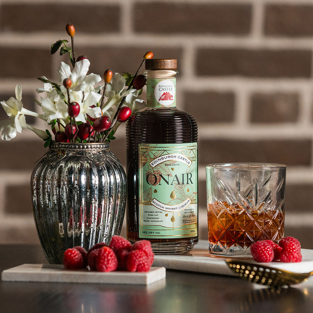 Bottle of Onair labelled liqueur surrounded by flowers, raspberries and a glass against a brick background.  - Image