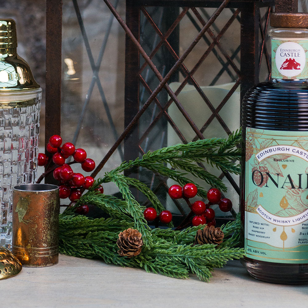 A bottle of Onair Whisky Liqueur and a smaller matching bottle, styled with a crystal shaker, brass jigger, and evergreen sprigs with red berries on a stone surface. - Image