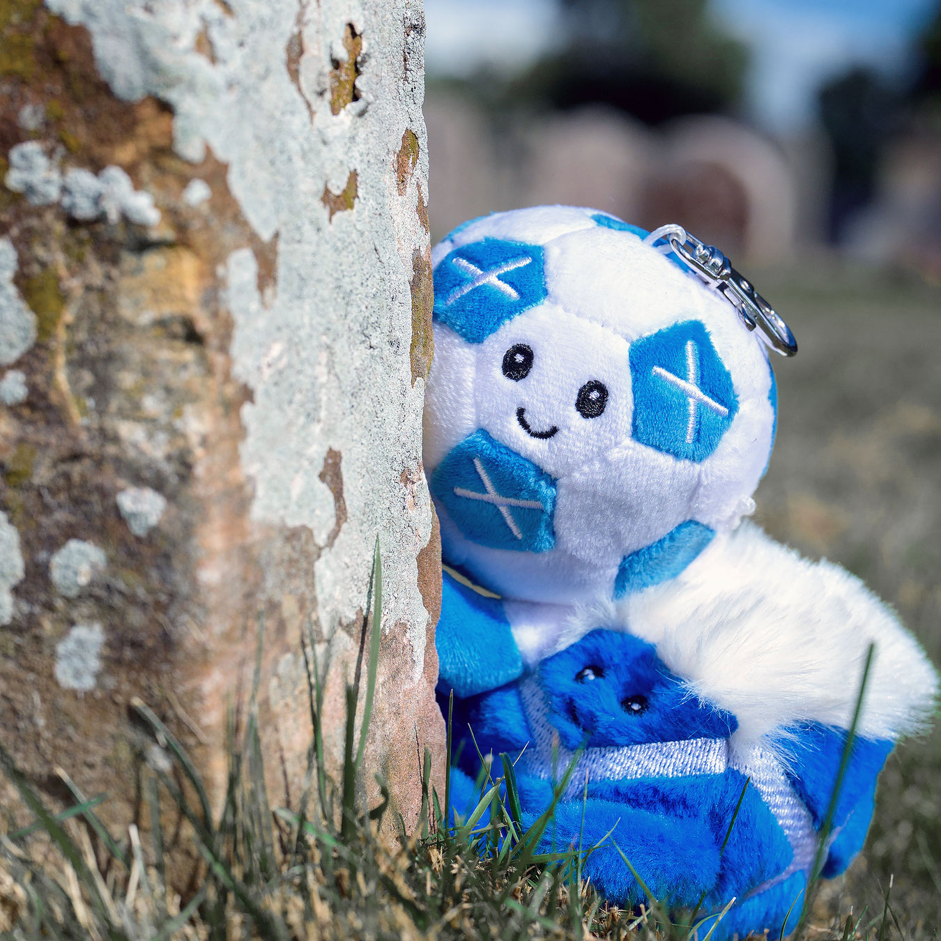 A blue and white soccer-themed plush keychain or small plush figure with embroidered facial features and a metal key ring, photographed outdoors beside a textured tree trunk in grass. - Image