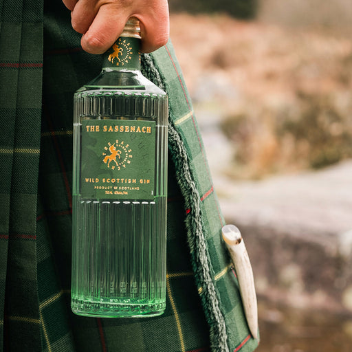 Bottle of Sassenach Wild Scottish Gin held by a person wearing a green kilt with a natural background. - Image