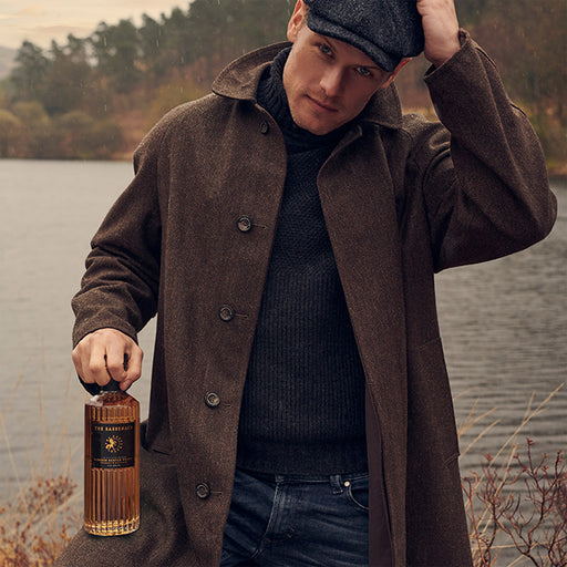 Man in a brown coat holding a bottle of The sassenach Whisky by a lake - Image