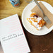 Book titled 'Who Will Be Remembered Here' next to a plate with dessert on a wooden table. - Image