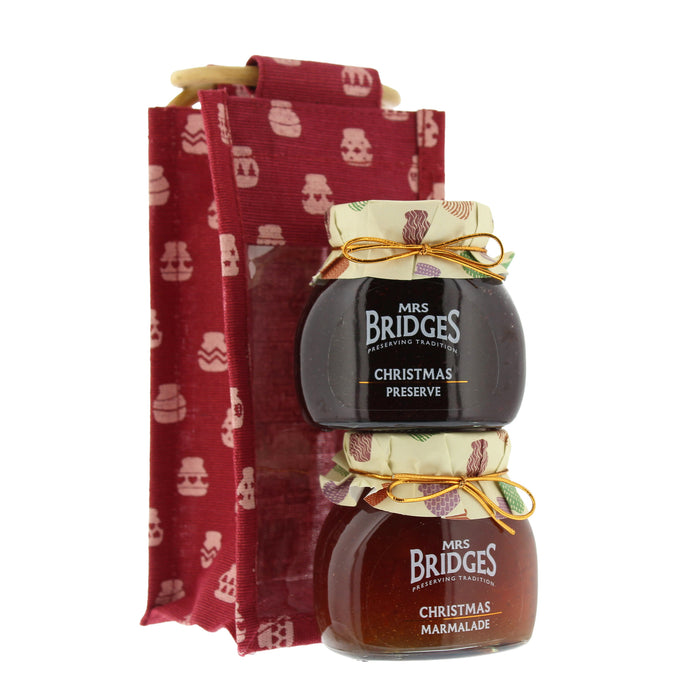 Two glass jars of Mrs. Bridges Christmas preserves stacked in front of a red fabric gift bag with white holiday-themed icons, on a white background. - Image