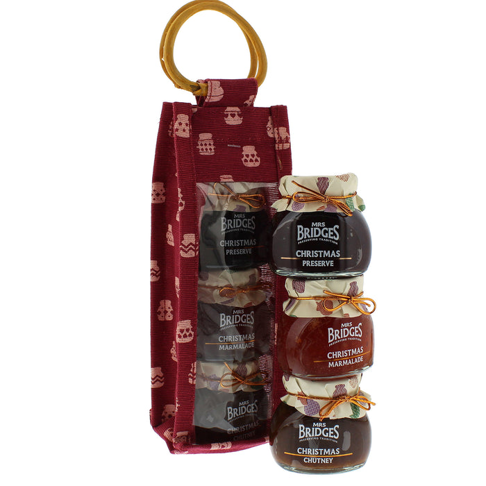 A gift set from Mrs Bridges featuring three glass jars of Christmas preserves stacked next to a red fabric pouch with a  ring handle.  - Image