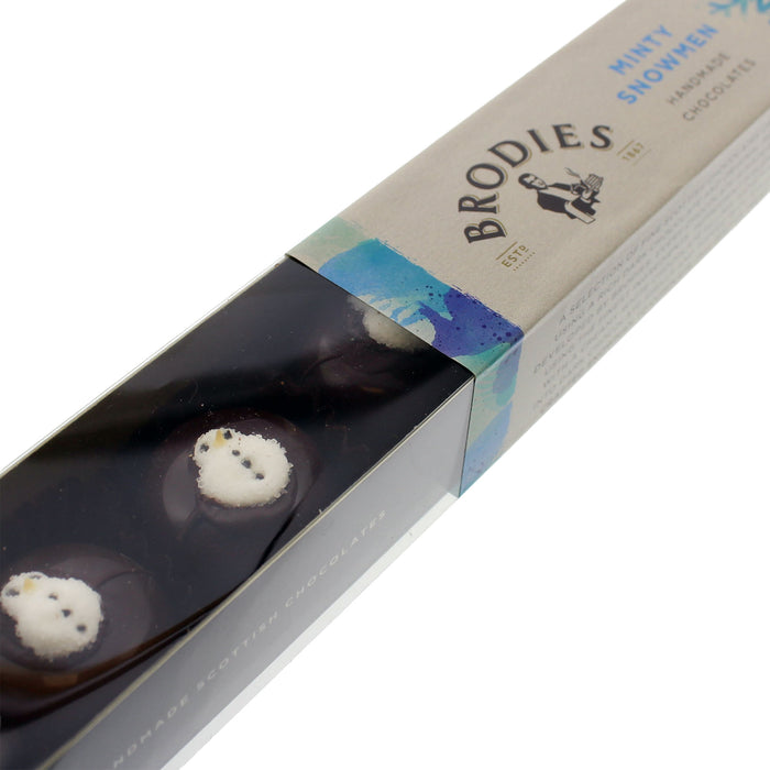 A long, angled view of a Brodies Minty Snowmen chocolate box. The packaging features a clear window showing two chocolate snowmen with white frosting and black eyes, set against a clean white background. - Image