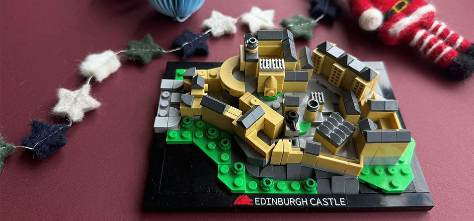 A custom-built Edinburgh Castle brick model on a black baseplate with green and gray bricks, photographed on a dark red surface with festive garlands in the background. - Image