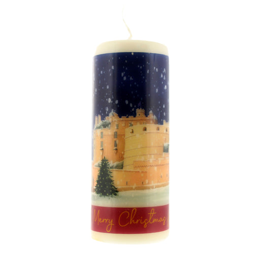 A cylindrical Christmas candle with a printed winter scene featuring a castle under a snowy night sky, with a red band at the bottom reading 'Merry Christmas' in gold script. The candle itself is off-white at the top and bottom. - Image