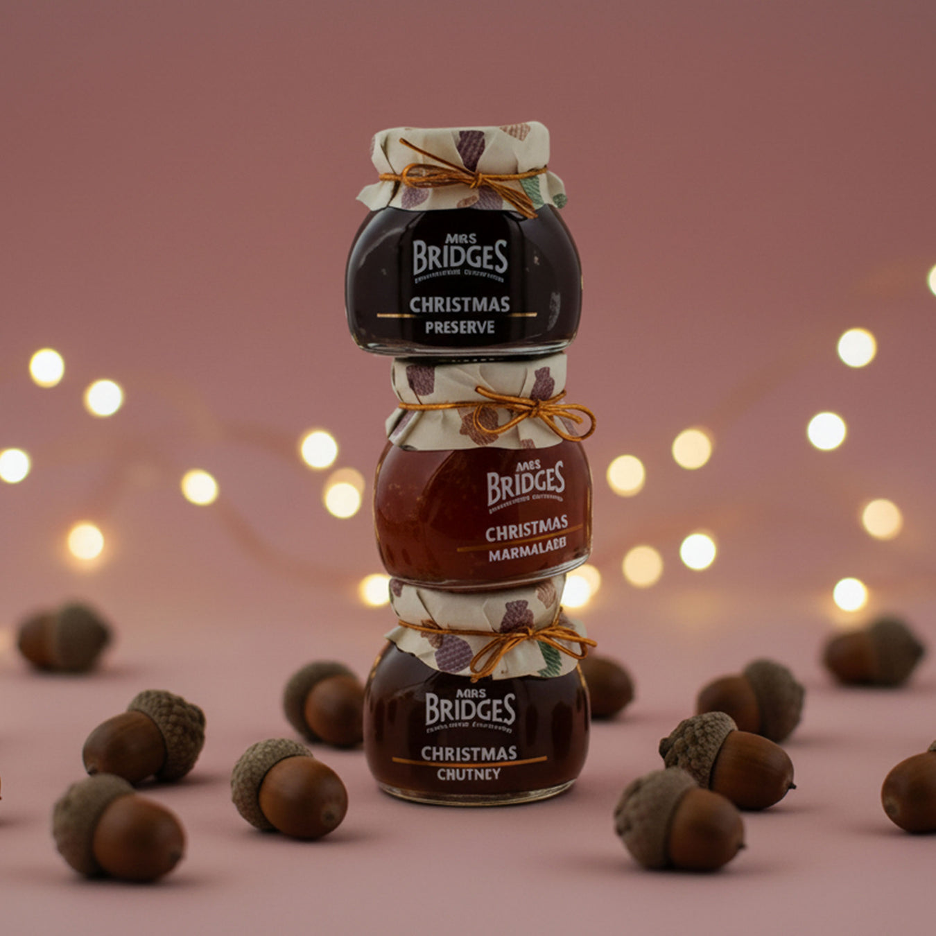 Three stacked jars of Mrs. Bridges Christmas preserves and marmalade tied together with orange string, set on a dusty pink surface with scattered acorns and warm fairy lights in the background. - Image