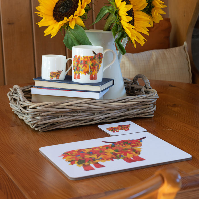 Autumn Highland Cow Mug shown with other items from the collection including placemat and coaster on a wooden table with sunflowers, books and wicker tray