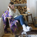 coorie tartan blanket shown on chair with model reading a book and logs in the background
